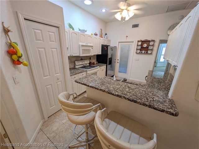 kitchen featuring kitchen peninsula, white appliances, dark stone countertops, white cabinets, and a breakfast bar area