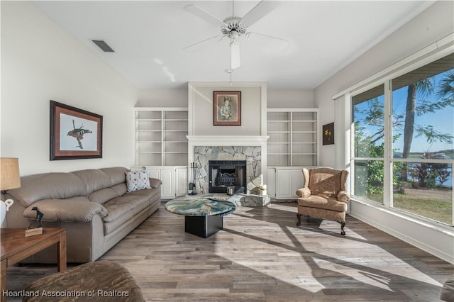living room featuring a stone fireplace, a ceiling fan, baseboards, wood finished floors, and visible vents