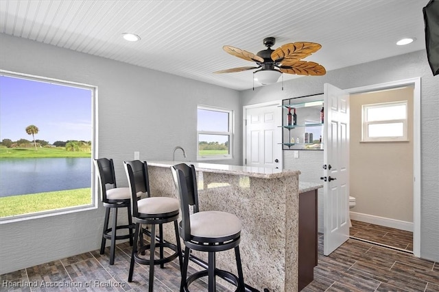 bar with ceiling fan, a water view, light stone countertops, and a wealth of natural light