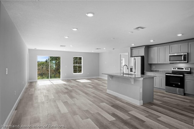 kitchen with gray cabinetry, light stone countertops, stainless steel appliances, light hardwood / wood-style flooring, and a kitchen island with sink