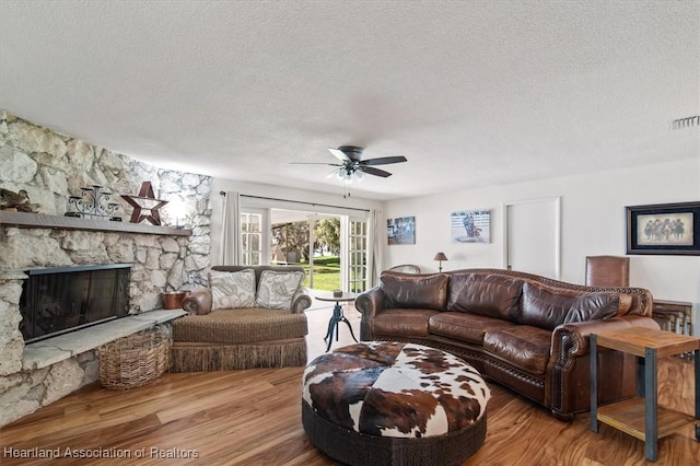 living room with ceiling fan, wood-type flooring, a stone fireplace, and a textured ceiling