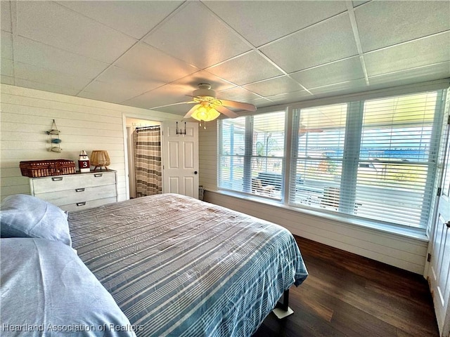 bedroom featuring ceiling fan, dark hardwood / wood-style flooring, a drop ceiling, and wooden walls