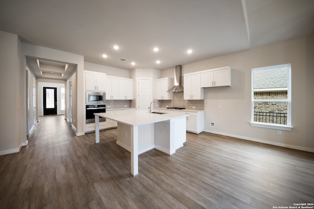 kitchen with white cabinetry, stainless steel appliances, wall chimney range hood, wood-type flooring, and a kitchen island with sink