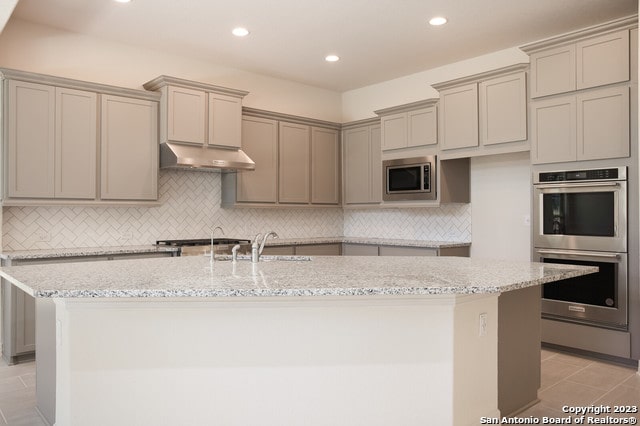 kitchen featuring gray cabinets, appliances with stainless steel finishes, light stone counters, and a kitchen island with sink