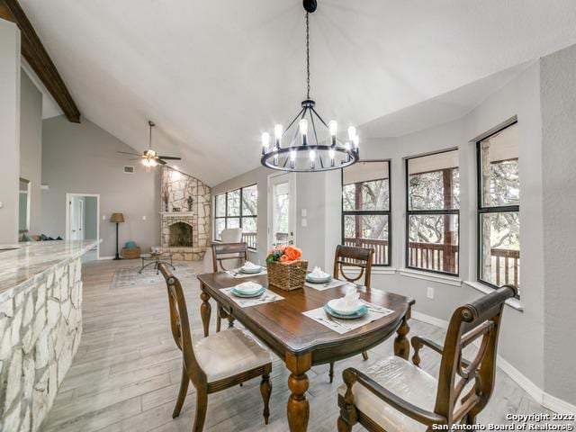 dining space featuring a stone fireplace, ceiling fan with notable chandelier, light wood-type flooring, and lofted ceiling with beams