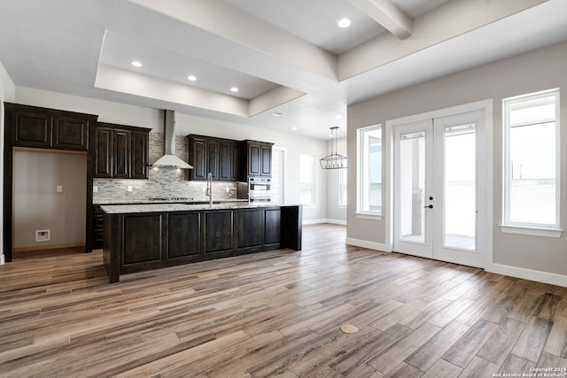 kitchen with french doors, wall chimney range hood, oven, an island with sink, and light hardwood / wood-style floors