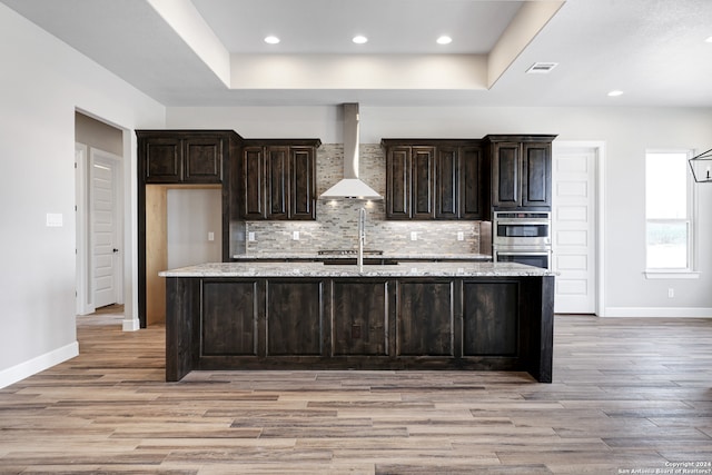 kitchen featuring light stone counters, stainless steel double oven, a kitchen island with sink, wall chimney range hood, and light hardwood / wood-style floors