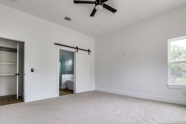 unfurnished bedroom featuring multiple windows, ceiling fan, a barn door, connected bathroom, and dark colored carpet