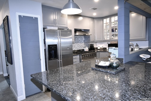 kitchen with hanging light fixtures, appliances with stainless steel finishes, dark stone counters, wall chimney range hood, and tasteful backsplash