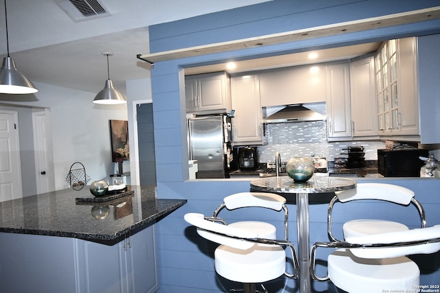 kitchen featuring stainless steel refrigerator, tasteful backsplash, wall chimney range hood, dark stone countertops, and decorative light fixtures