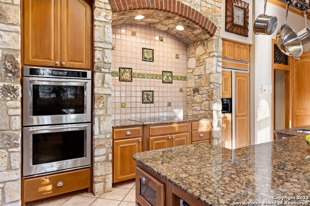 kitchen with tile walls, built in appliances, light tile floors, dark stone counters, and tasteful backsplash