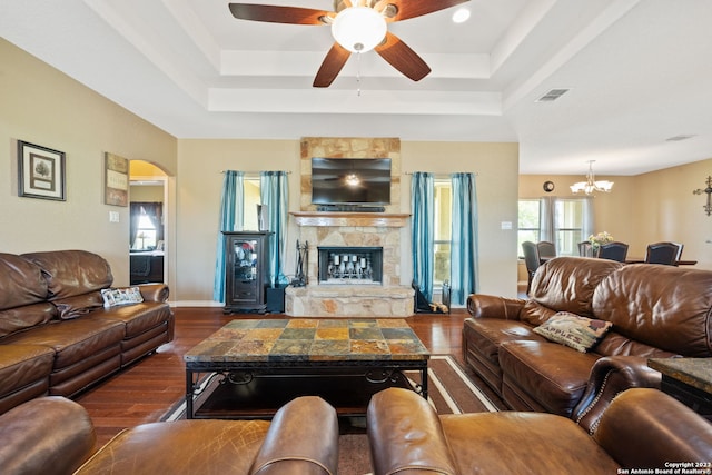 living room with a raised ceiling, dark wood-type flooring, a stone fireplace, and ceiling fan with notable chandelier