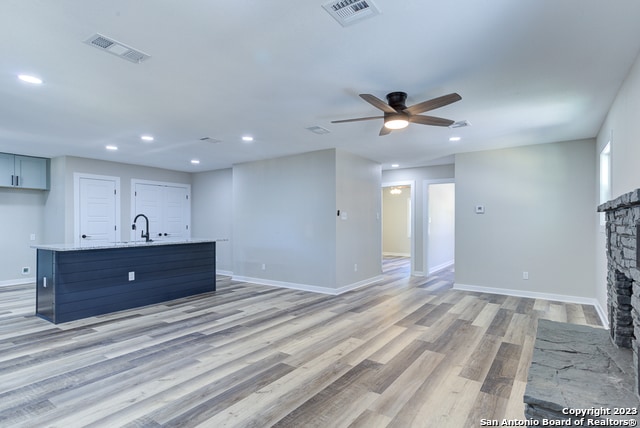 unfurnished living room with sink, ceiling fan, a stone fireplace, and light wood-type flooring