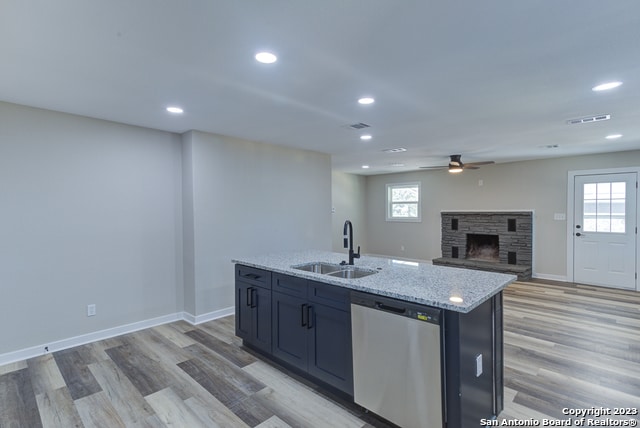 kitchen with ceiling fan, an island with sink, light hardwood / wood-style flooring, dishwasher, and a stone fireplace