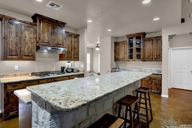 kitchen featuring light stone countertops, ceiling fan, stainless steel gas stovetop, a breakfast bar, and dark tile floors