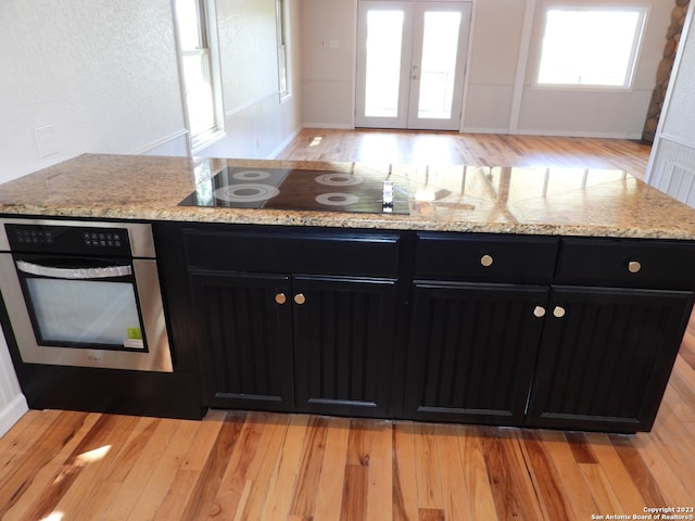 kitchen with light wood-style flooring, light stone countertops, black electric cooktop, dark cabinetry, and stainless steel oven