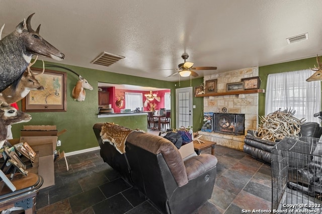 tiled living room with a textured ceiling, a stone fireplace, ceiling fan with notable chandelier, and a healthy amount of sunlight