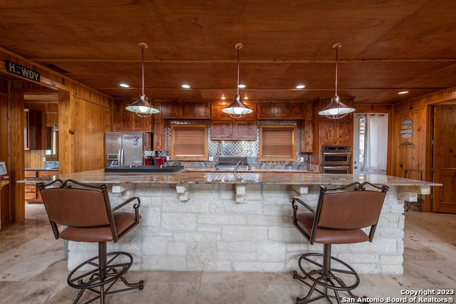 kitchen featuring hanging light fixtures, wooden walls, stainless steel fridge, and a kitchen bar