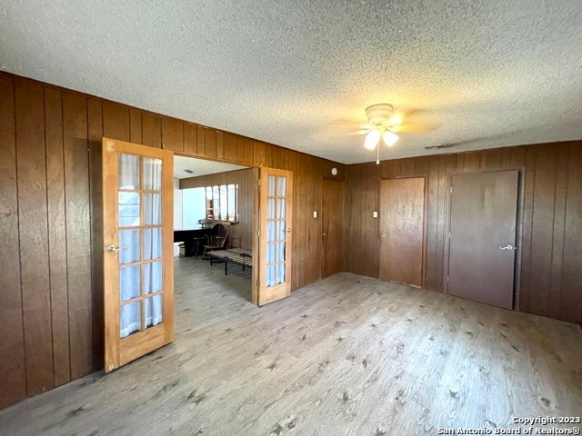 unfurnished room featuring french doors, light hardwood / wood-style flooring, wooden walls, ceiling fan, and a textured ceiling