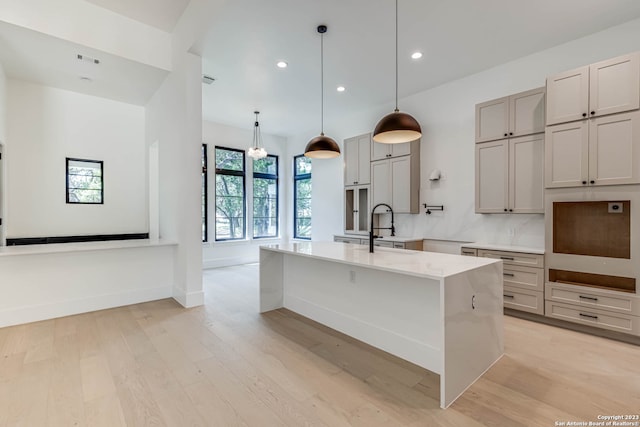 kitchen featuring light hardwood / wood-style floors, a kitchen island with sink, light stone countertops, backsplash, and hanging light fixtures