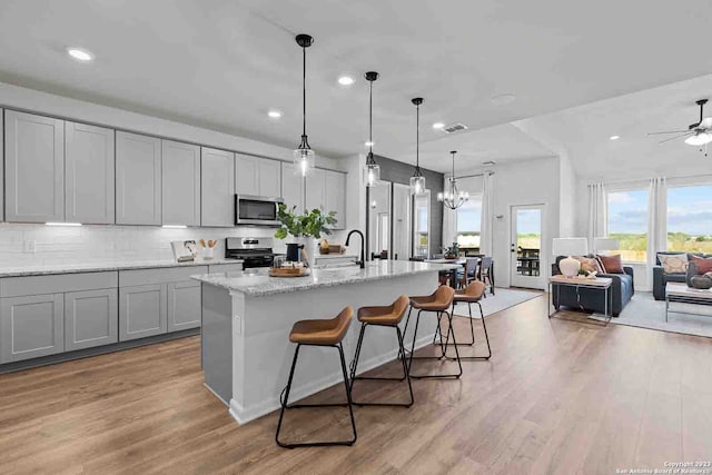 kitchen with ceiling fan with notable chandelier, gray cabinets, appliances with stainless steel finishes, a kitchen island with sink, and light wood-type flooring