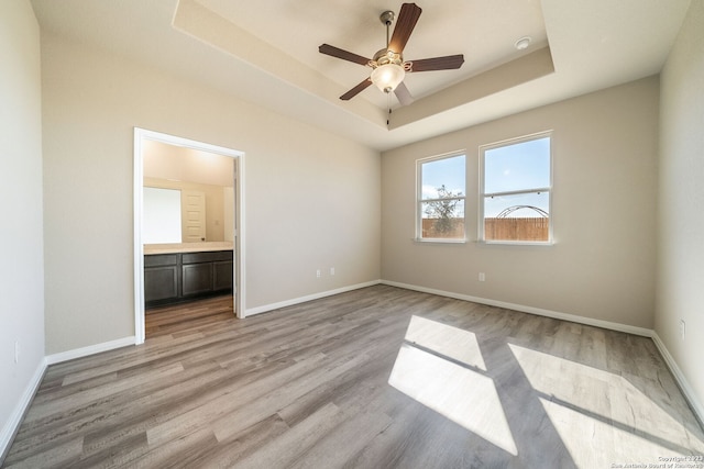 unfurnished bedroom with light wood-type flooring, connected bathroom, a tray ceiling, and ceiling fan