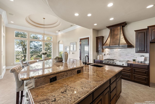 kitchen with a tray ceiling, a kitchen breakfast bar, a center island with sink, custom range hood, and light stone counters