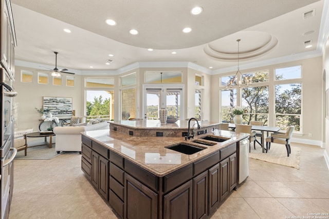 kitchen with hanging light fixtures, dishwasher, ceiling fan with notable chandelier, a center island with sink, and a tray ceiling