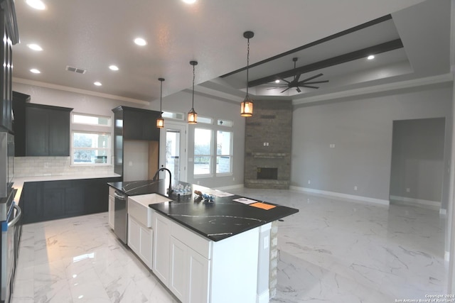 kitchen featuring a stone fireplace, white cabinetry, ornamental molding, a raised ceiling, and a center island with sink