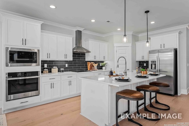 kitchen with white cabinetry, wall chimney exhaust hood, stainless steel appliances, light hardwood / wood-style flooring, and a kitchen island with sink