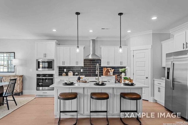 kitchen featuring wall chimney exhaust hood, stainless steel appliances, a kitchen island with sink, and decorative light fixtures