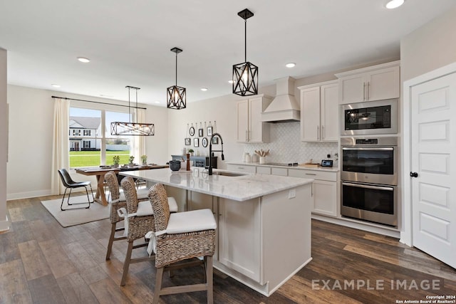 kitchen with custom range hood, double oven, a kitchen island with sink, sink, and white cabinets