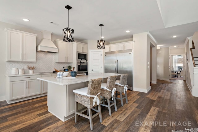 kitchen with premium range hood, white cabinets, a center island with sink, hanging light fixtures, and built in appliances