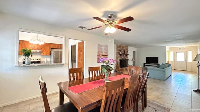 tiled dining space featuring ceiling fan with notable chandelier and a brick fireplace