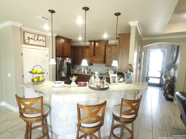 kitchen featuring hanging light fixtures, light stone countertops, stainless steel fridge, ornamental molding, and a kitchen breakfast bar