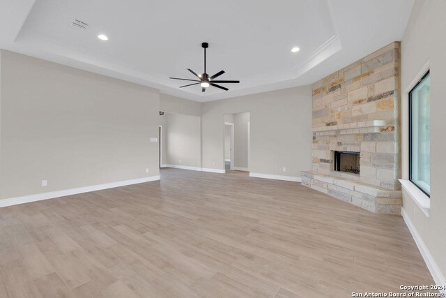 unfurnished living room with ceiling fan, a stone fireplace, light wood-type flooring, and a tray ceiling