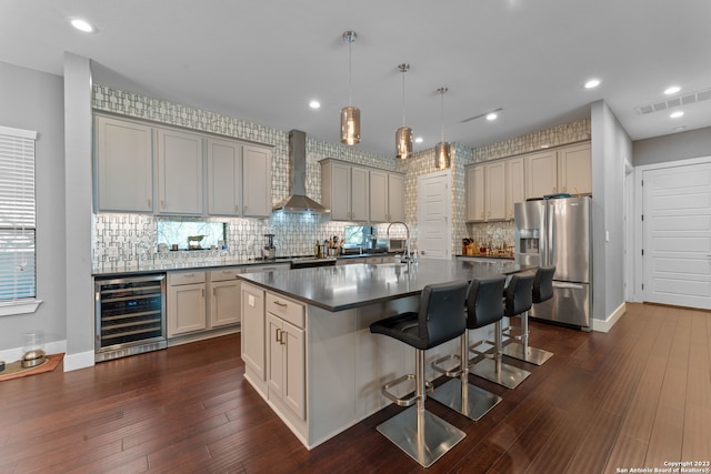 kitchen featuring stainless steel fridge, a kitchen breakfast bar, a center island with sink, beverage cooler, and wall chimney range hood