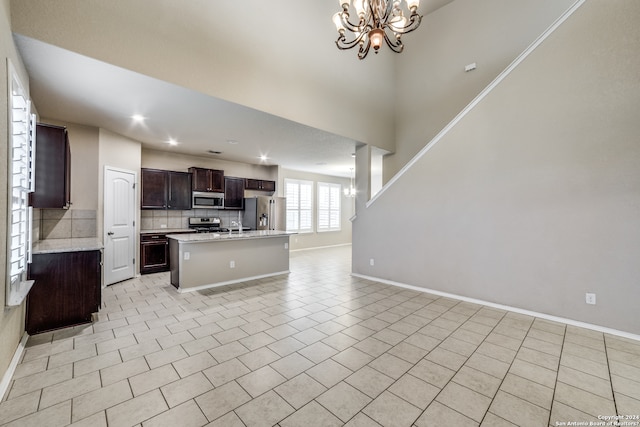 kitchen with appliances with stainless steel finishes, backsplash, a center island with sink, light tile flooring, and an inviting chandelier