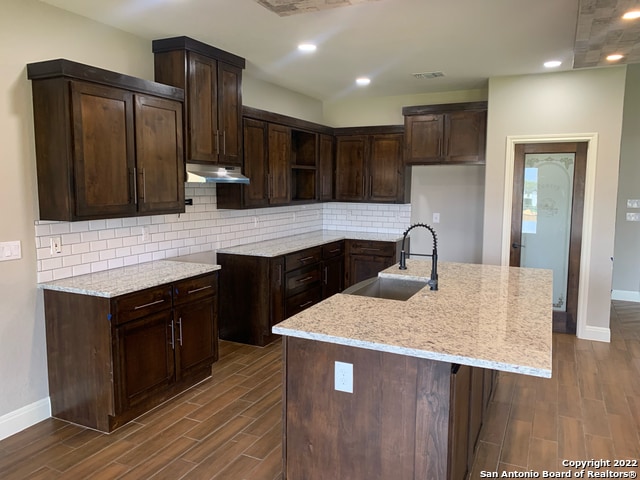 kitchen with light stone counters, tasteful backsplash, dark brown cabinets, a kitchen island with sink, and sink