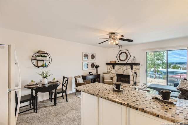 kitchen featuring light colored carpet, white fridge, ceiling fan, a stone fireplace, and white cabinets