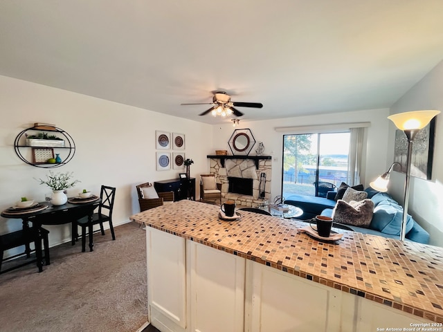 kitchen with white cabinets, dark carpet, a stone fireplace, and ceiling fan