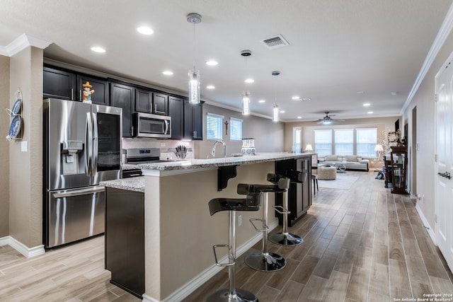 kitchen with stainless steel appliances, light stone countertops, ceiling fan, ornamental molding, and a kitchen island with sink