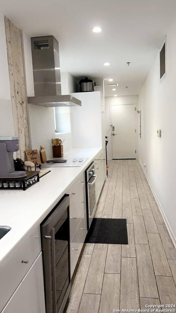 kitchen featuring beverage cooler, oven, light hardwood / wood-style flooring, wall chimney exhaust hood, and white cabinets