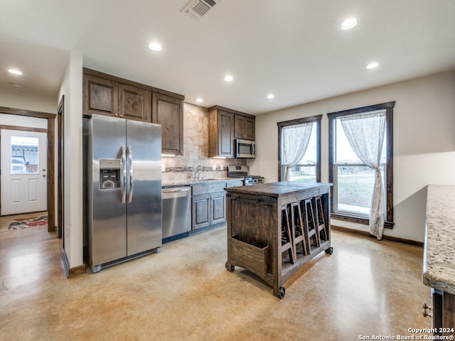 kitchen featuring appliances with stainless steel finishes, light stone counters, a kitchen island, tasteful backsplash, and dark brown cabinetry