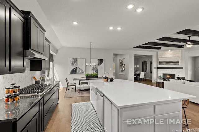 kitchen featuring light hardwood / wood-style floors, a stone fireplace, an island with sink, ceiling fan with notable chandelier, and beam ceiling