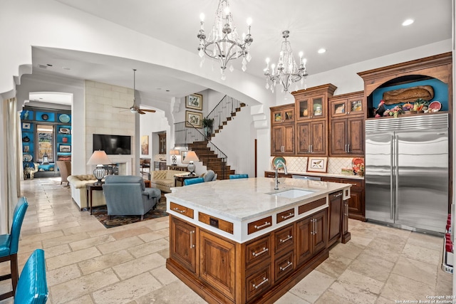 kitchen with ceiling fan with notable chandelier, a kitchen island with sink, stainless steel built in refrigerator, light tile patterned floors, and sink