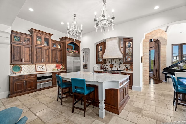 kitchen featuring premium range hood, decorative backsplash, built in refrigerator, an island with sink, and sink