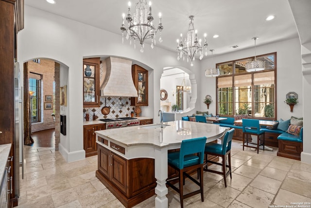 kitchen featuring decorative backsplash, custom range hood, pendant lighting, light tile patterned floors, and an island with sink