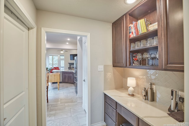 bar with built in desk, light stone counters, backsplash, ceiling fan, and dark brown cabinetry