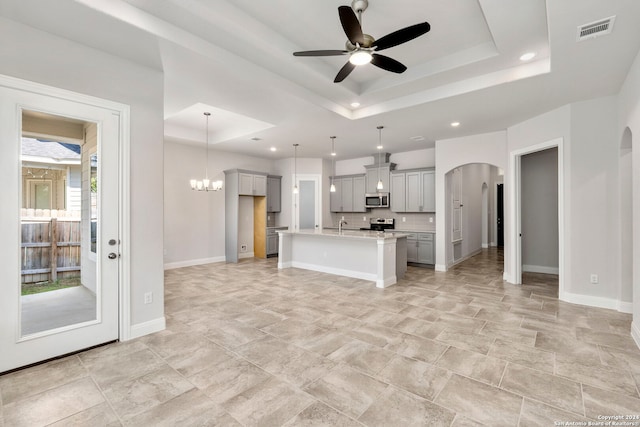 kitchen with ceiling fan with notable chandelier, appliances with stainless steel finishes, a center island with sink, a raised ceiling, and gray cabinets